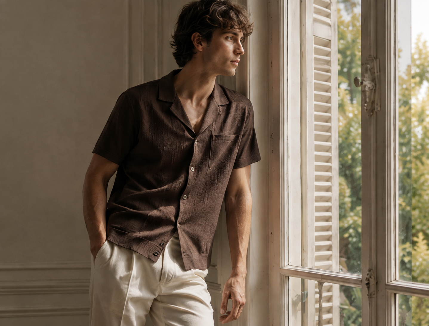 Man in a brown textured shirt and ivory trousers beside a window